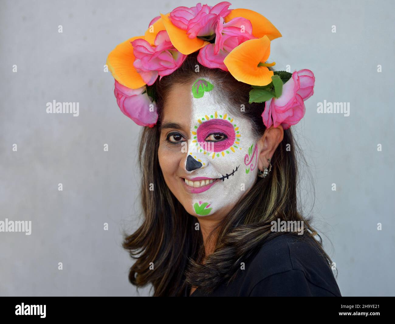 Donna messicana con fiori colorati in capelli dipinge la metà sinistra del suo volto con il viso Catrina make-up il giorno dei morti e sorride allo spettatore. Foto Stock