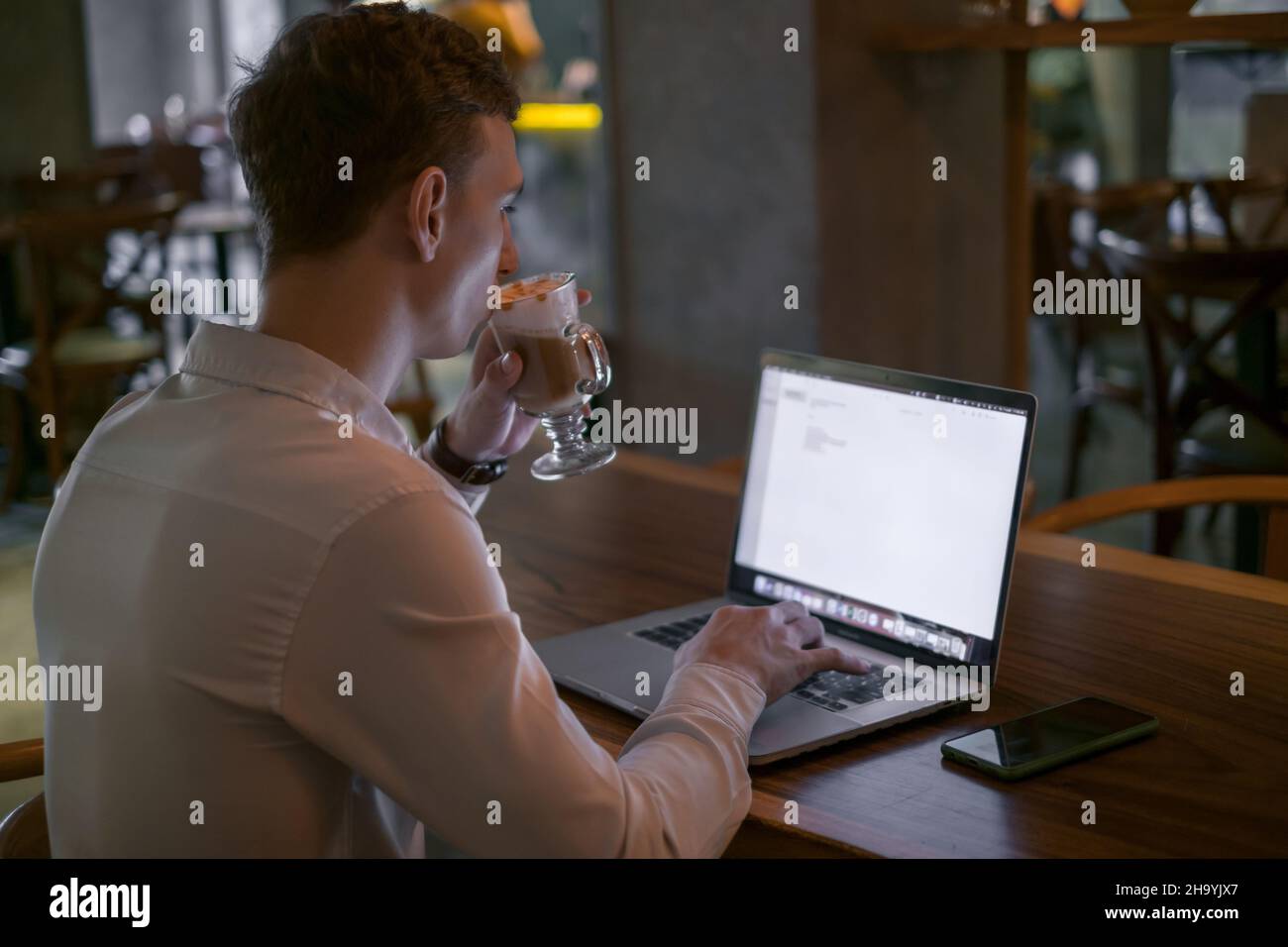 Giovane uomo d'affari caucasico che naviga in rete su un computer portatile mentre beve caffè in un bar. Bell'uomo che lavora in un bar con un computer portatile. Digitazione sulla tastiera Foto Stock