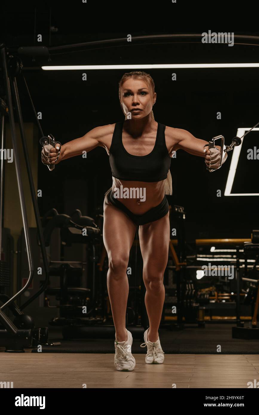 Una donna sportiva con capelli biondi sta facendo un allenamento al petto sulla macchina del cavo in una palestra. Una ragazza sta addestrando i suoi muscoli pettorali. Foto Stock