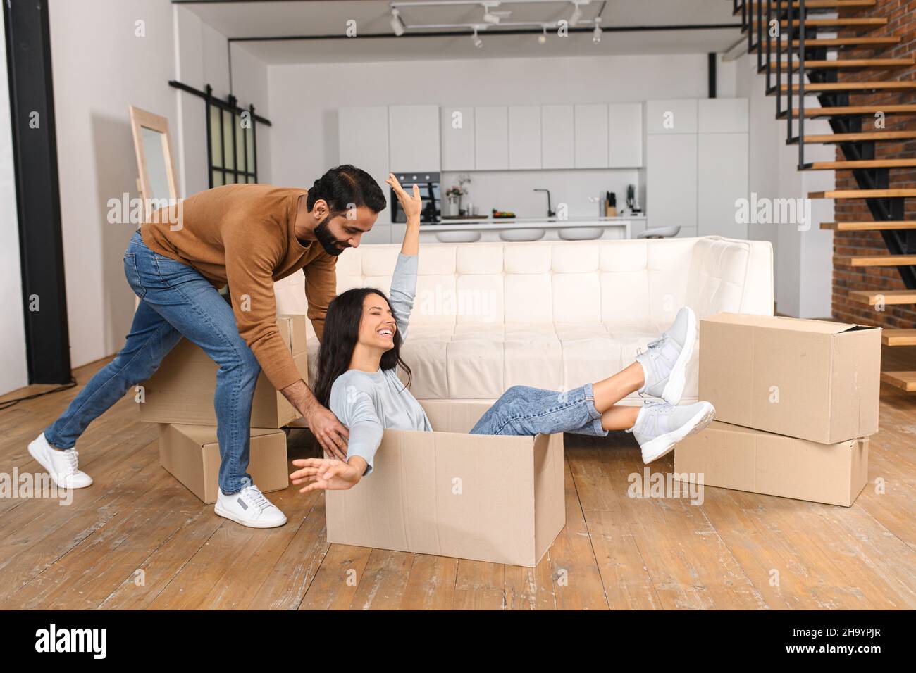 Felice coppia multirazziale prima volta acquirenti casa che hanno divertimento mentre disimballaggio effetti personali su giorno in movimento. Eccitata moglie che cavalcava in scatola di cartone mentre suo marito lo spingeva in un nuovo appartamento Foto Stock