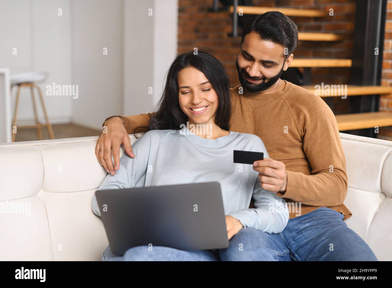 Coppia indiana allegra che guarda allo schermo del laptop e controlla il numero della carta di credito per effettuare la transazione online. Sposi sorridenti che hanno shopping in linea. Trasferimento di denaro, concetto di online banking Foto Stock