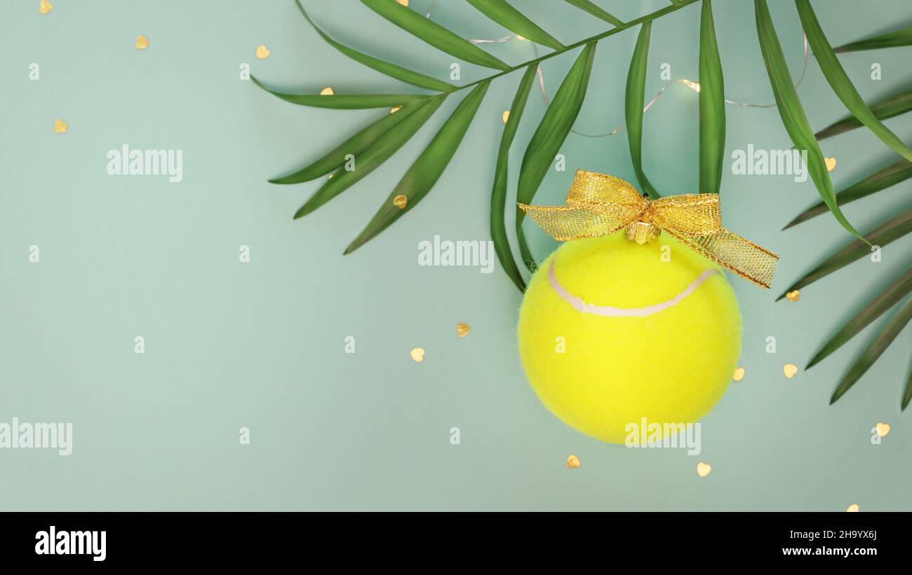 Tennis, composizione vacanze invernali con palla da tennis gialla sotto forma di palla di Capodanno, foglie di palma e confetti su sfondo blu con copia Foto Stock