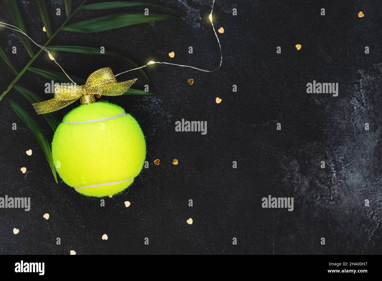 Tennis, composizione vacanze invernali con palla da tennis gialla sotto forma di palla di Capodanno, foglie di palma e confetti su sfondo nero con copia Foto Stock