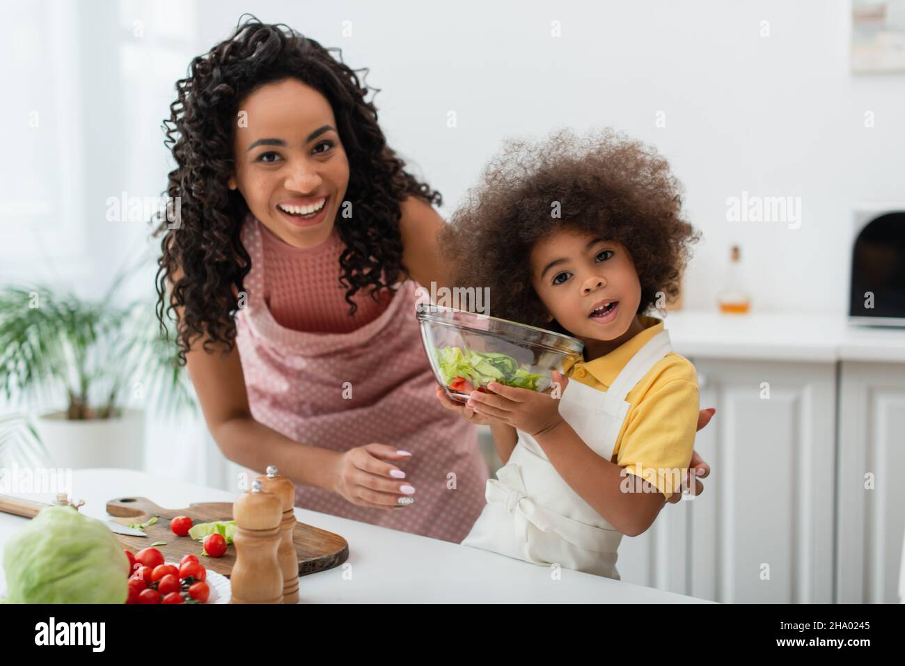 Sorridente donna americana africana guardando la macchina fotografica vicino alla figlia con ciotola di insalata a casa Foto Stock