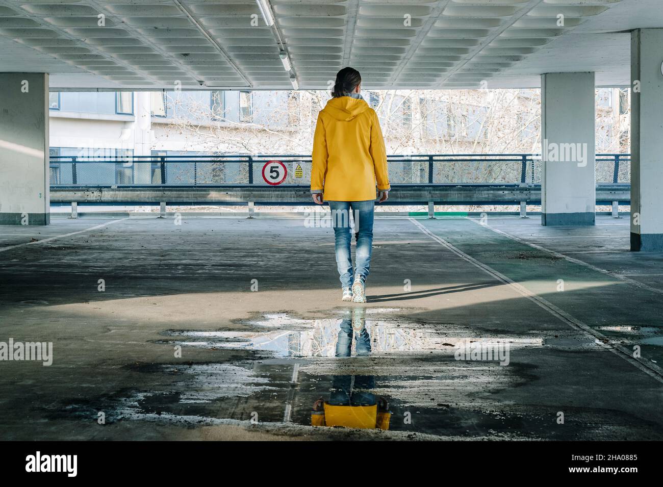 Vista posteriore di una donna che si allontana in un parcheggio in cemento con una giacca gialla. Foto Stock