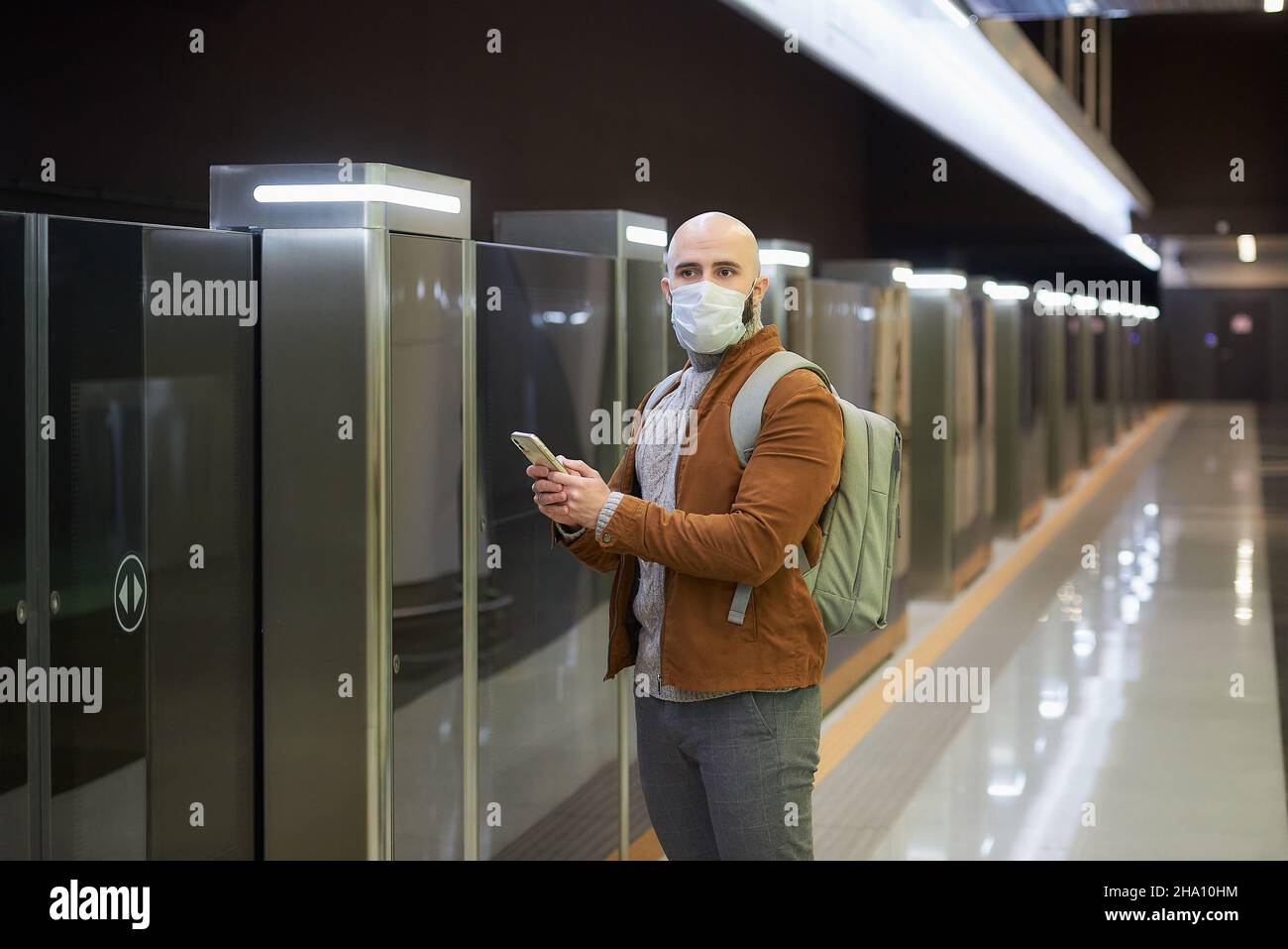 Un uomo in una maschera medica sta tenendo un telefono e fissando al lato mentre aspetta un treno alla piattaforma della metropolitana. Un ragazzo calvo in un mas chirurgico Foto Stock