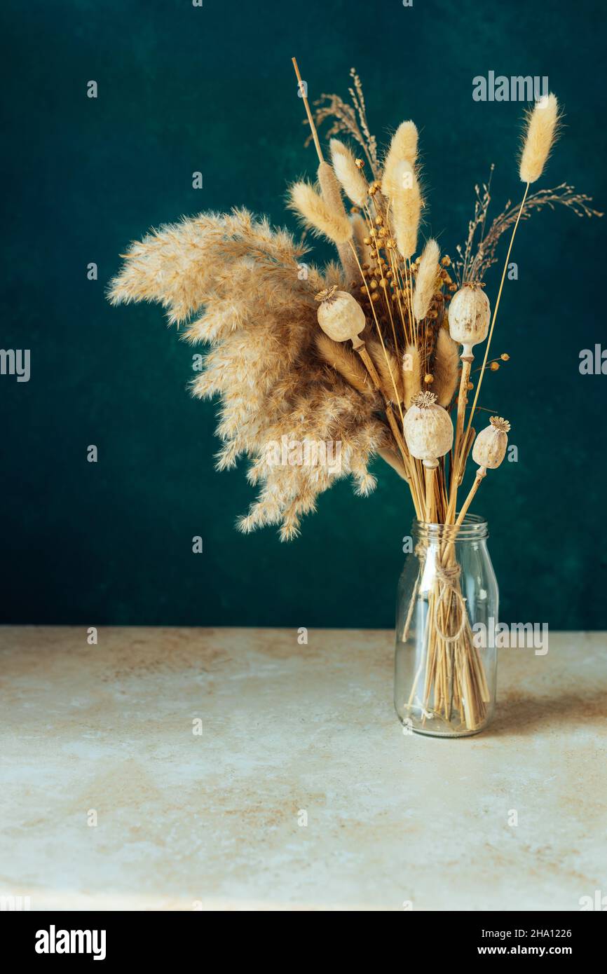 Bouquet di fiori secchi beige in vaso di vetro su sfondo verde-blu. Foto Stock