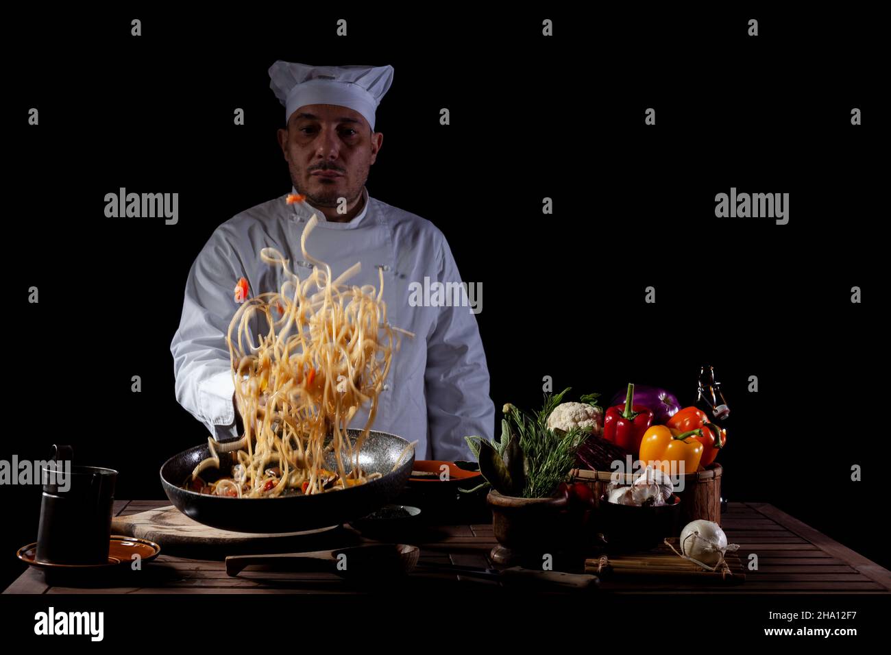 Cuoco maschile in uniforme bianca che tiene una padella, soffriggete gli spaghetti con verdure fresche che volano nell'aria prima di servire mentre lavorate in un ristorante Foto Stock