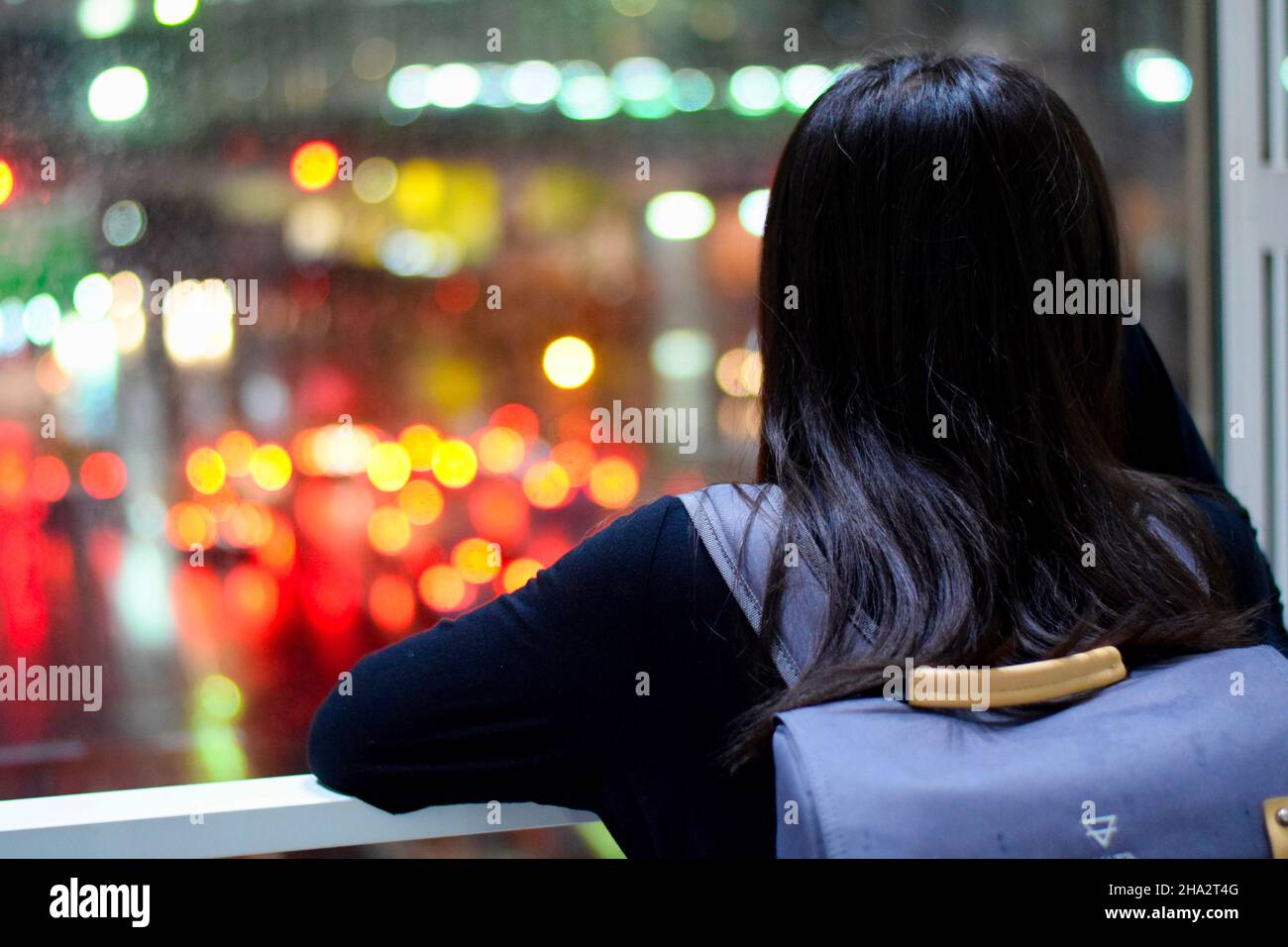 Vista posteriore di una ragazza che guarda verso l'esterno una finestra alle luci blurry delle automobili nella pioggia Foto Stock