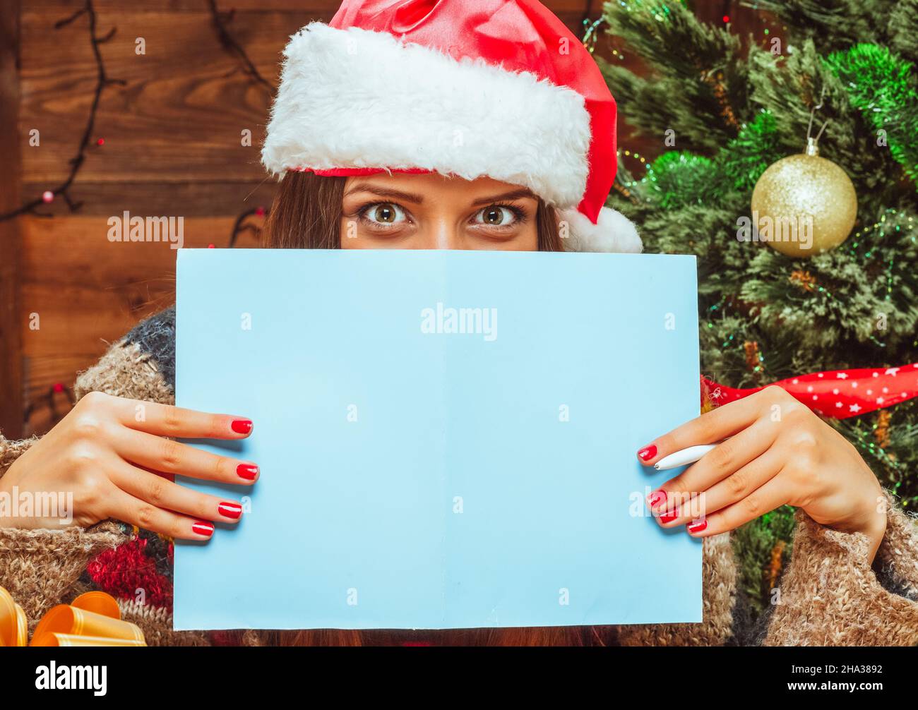 Modello caucasico femmina in un cappello Santa coperto la parte inferiore del suo viso con un foglio di carta bluastro, ha una penna in mano. Lettera a Babbo Natale. Lettera a Padre Frost, una lista di desideri, un piano per il prossimo anno nuovo. Primo piano. Legno sfondo. Foto di alta qualità Foto Stock
