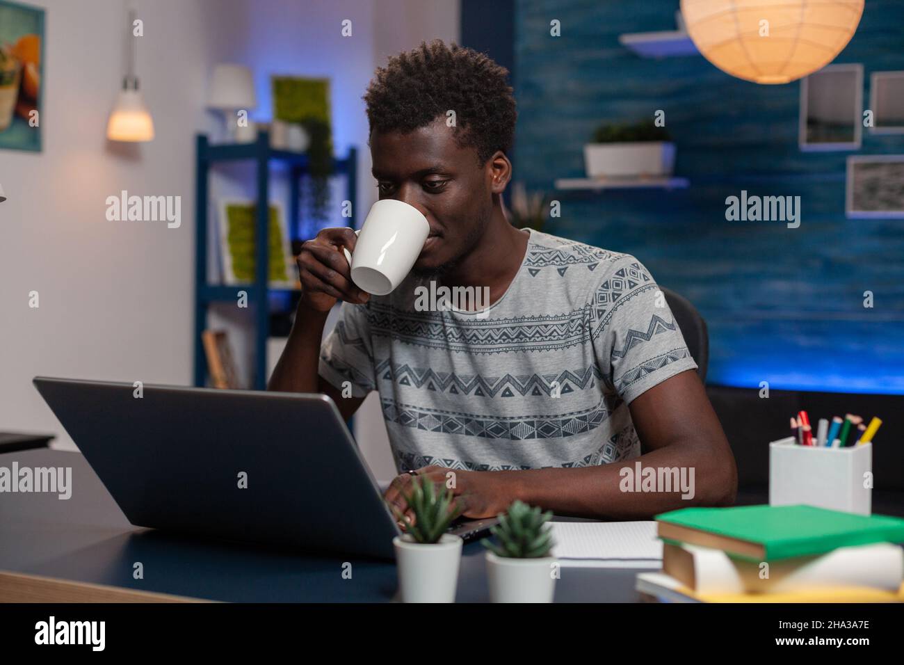 Ritratto di giovane imprenditore afroamericano seduto alla scrivania digitando idee di marketing analisi del grafico finanziario su computer portatile. Il dipendente beve caffè mentre lavora lontano da casa Foto Stock