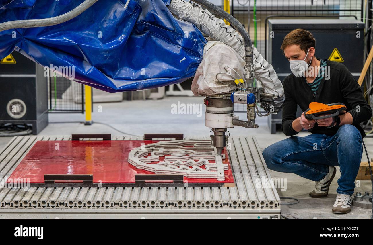 10 dicembre 2021, Sassonia, Chemnitz: Enrico Rudolph dell'area di ricerca "strutture leggere in ingegneria civile" dell'Università di tecnologia Chemnitz segue la produzione di un arco in calcestruzzo flyweight utilizzando un processo di stampa automatizzato 3D. Gli scienziati dell'Institute for Lightweight Structures presso la Chemnitz University of Technology ricercano tecnologie automatizzate di produzione di additivi per cinque anni. Recentemente, hanno presentato il primo contrafforte volante in calcestruzzo stampato a 3D° del mondo. Una speciale formula di cemento rinforzato con fibre viene applicata in diversi strati da un robot. T Foto Stock