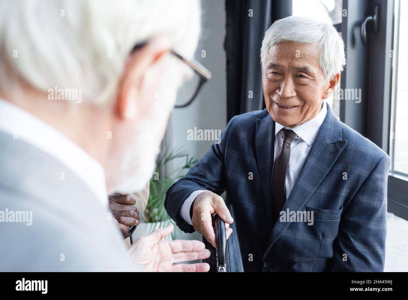 uomo d'affari asiatico anziano sorridente mentre dà la valigetta al partner d'affari blurred Foto Stock