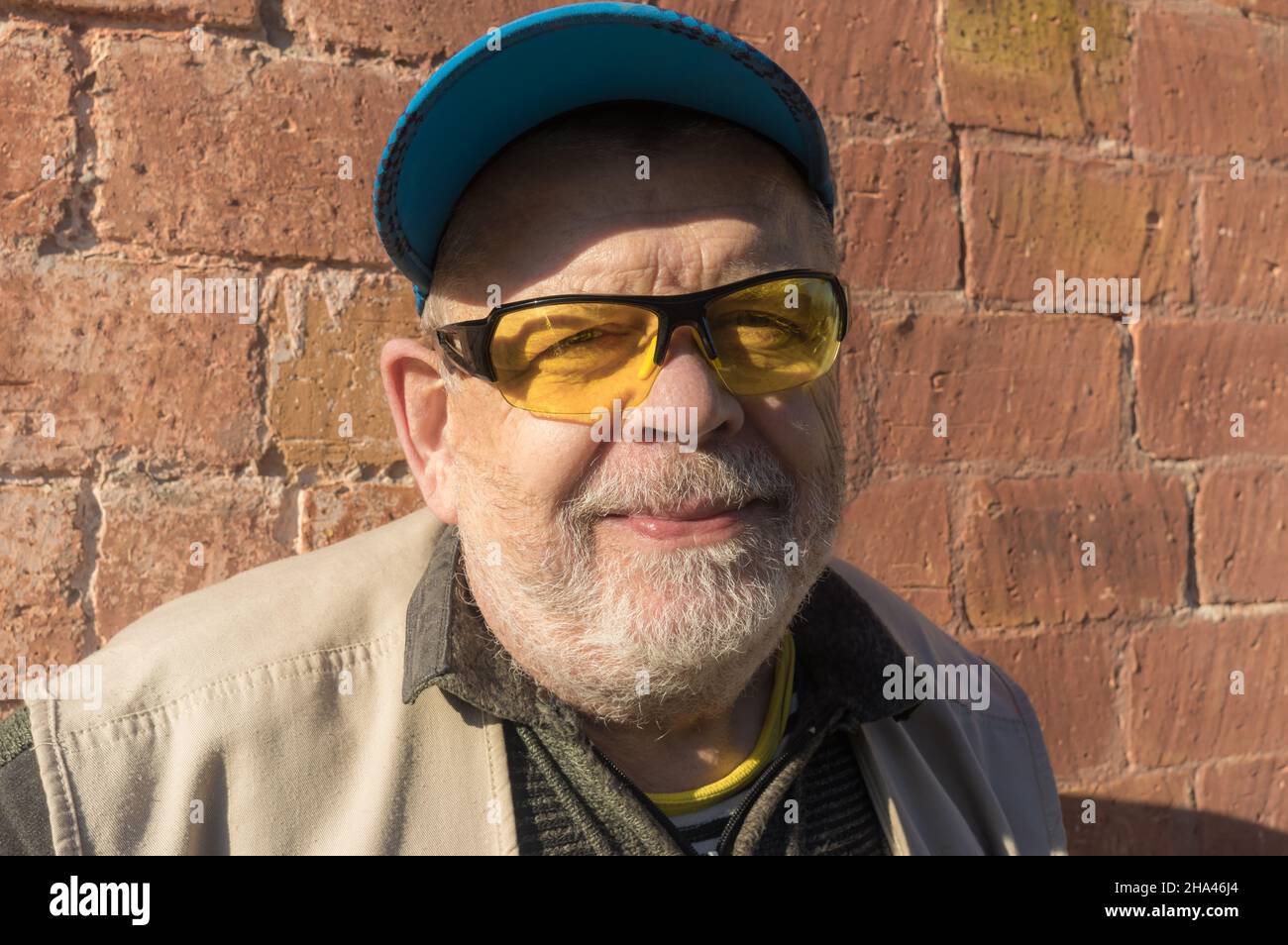Ritratto all'aperto di uomo anziano caucasico sorridente con cappuccio blu e occhiali da sole gialli mentre si alza contro una parete di mattoni pieni Foto Stock