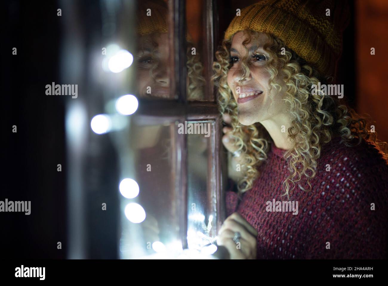 felice giovane donna adulta guardare fuori dalle finestre a casa di notte. le donne con capelli ricci e cappello a maglia godono al coperto nella stagione invernale. luci di festa e riflessione sul vetro Foto Stock