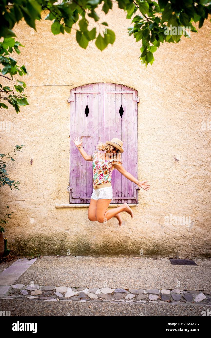 spensierate bella donna in cappello che salta a metà strada di fronte alla vecchia finestra di legno chiusa della parete testurizzata dell'edificio. estatica donna hipster che salta in aria all'aperto durante la vacanza Foto Stock