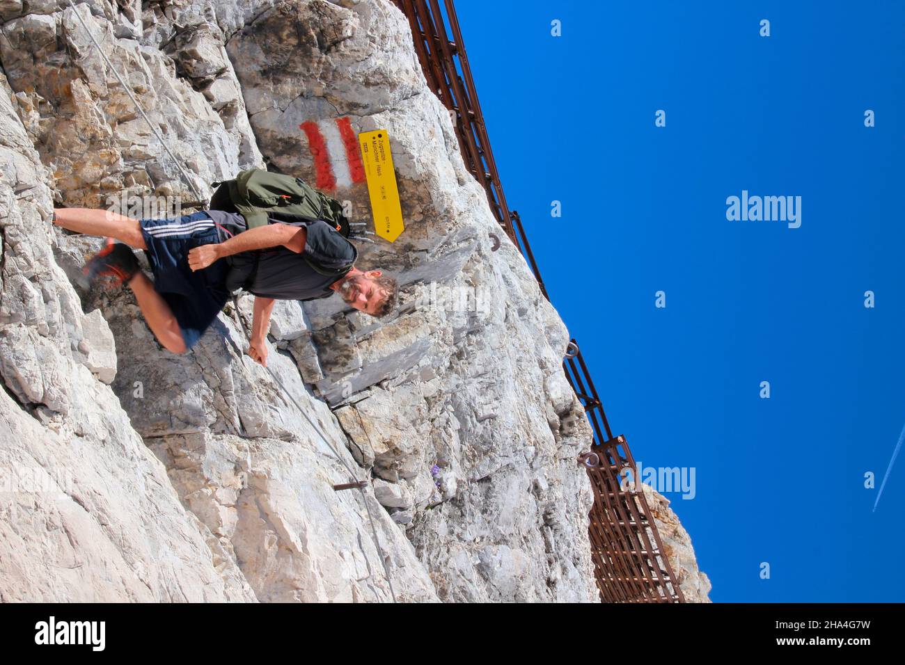 uomo di mezza età sulla via ferrata a zugspitze, guardare lateralmente, cartello, wetterstein montagne blu cielo, zaino, garmisch-partenkirchen, loisachtal, alta baviera, baviera, germania meridionale, germania, europa, Foto Stock