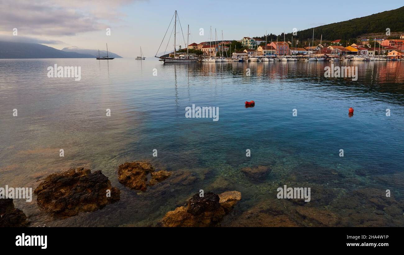 grecia,isole greche,isole ionie,cefalonia,fiskardo,umore del mattino,cielo parzialmente nuvoloso,vista da est sul bacino del porto a fiskardo. in primo piano un mare verde-blu con rocce,boe rosse a metà distanza,una grande barca a vela e altre barche a vela sono all'ancora Foto Stock