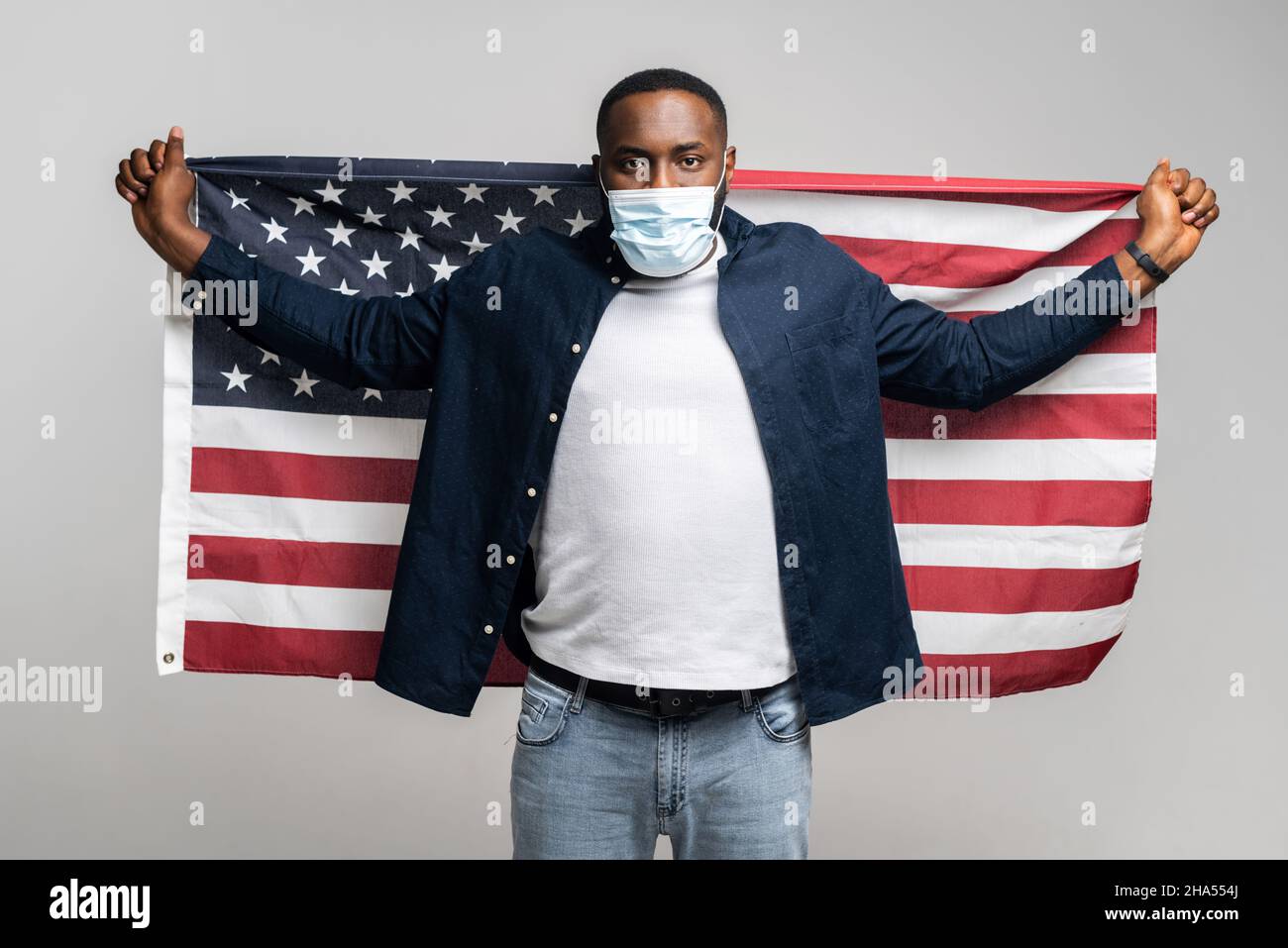 Giovane uomo afro-americano combattente per i diritti in maschera medica di protezione tenendo aperta la bandiera degli Stati Uniti dietro la schiena, sorridente e allegro ragazzo protestante contro il razzismo, isolato sul muro grigio Foto Stock