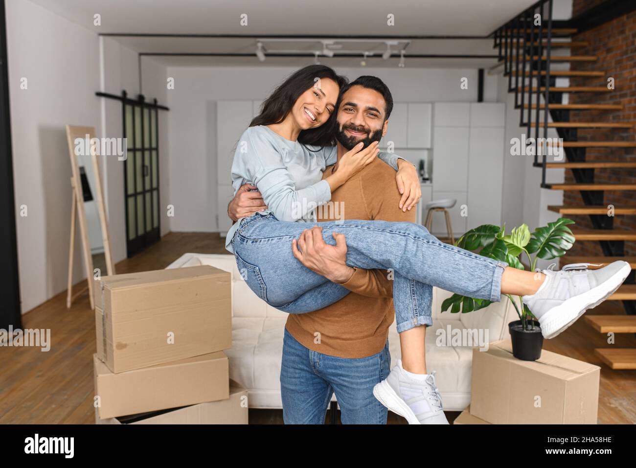 Marito pakistano che solleva e abbraccia sua moglie mentre festeggia il passaggio in un nuovo appartamento. Casa nostra, felice di passare a nuovo concetto di casa Foto Stock