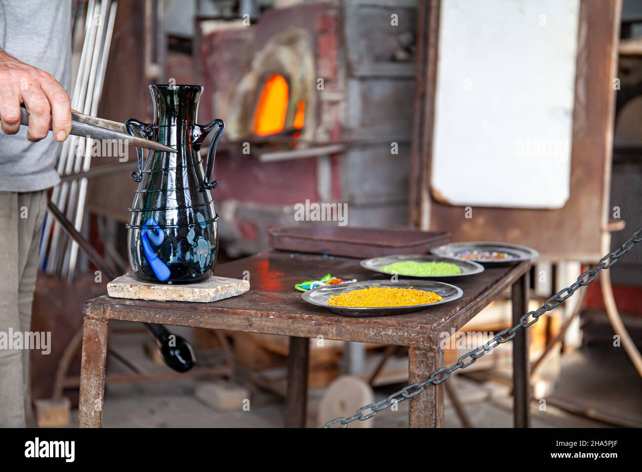 Un artigiano veneziano del vetro sull'isola di Murano sta lavorando su un vaso di vetro soffiato su un tavolo da lavoro in ferro con un'asta metallica. Venezia, Italia. Foto Stock