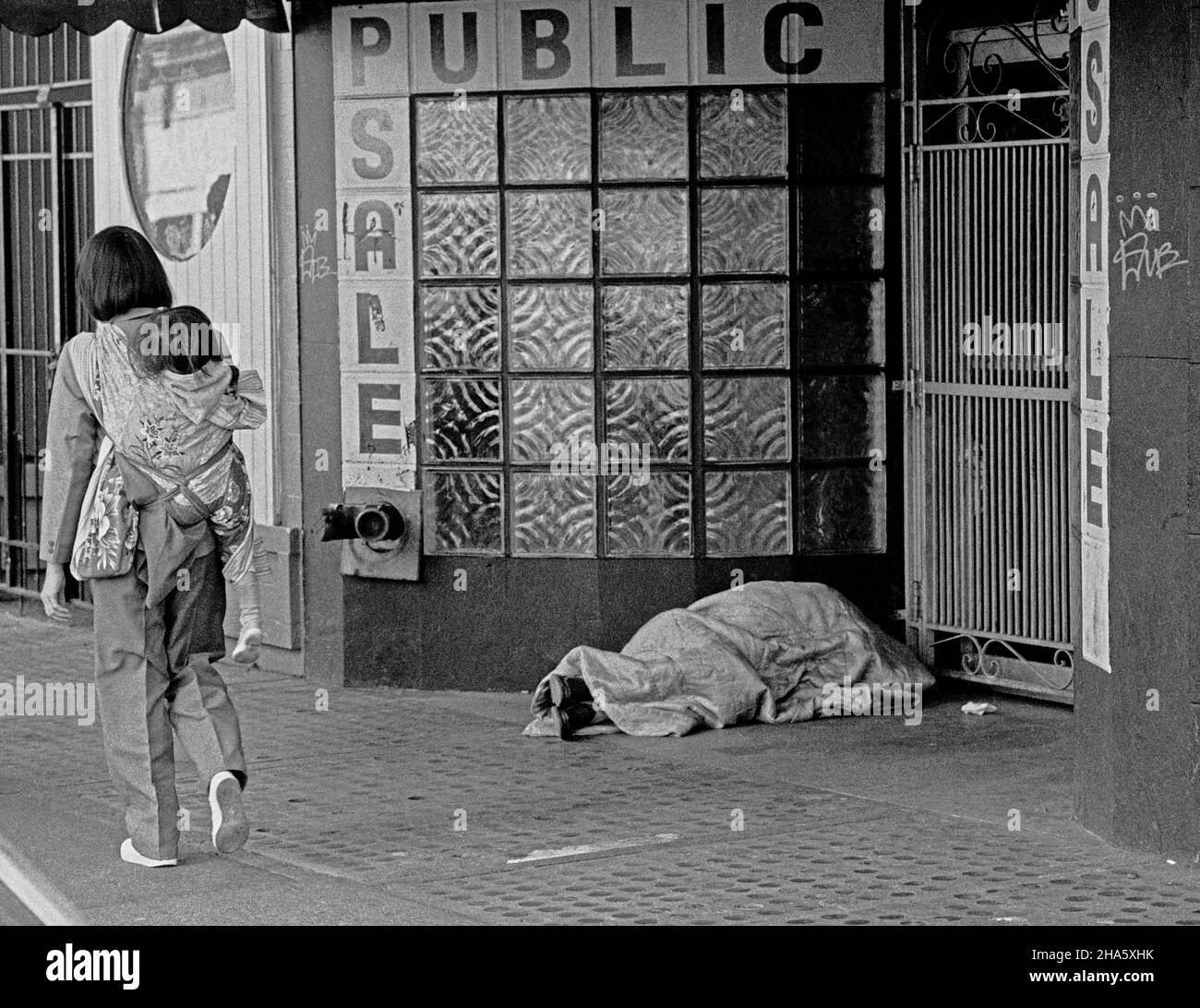 La donna che porta un bambino sulla schiena cammina accanto a una persona che dorme in una porta su un marciapiede di San Francisco nel 1980s Foto Stock