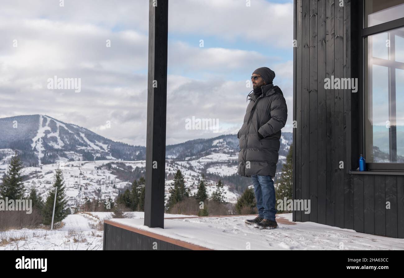Uomo in piedi sulla terrazza della casa e guardando le montagne innevate Foto Stock