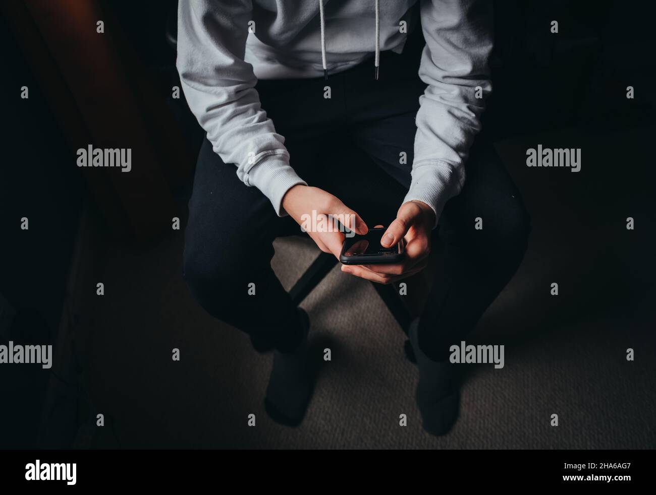 Primo piano di ragazzo teen in maglione bianco SMS sul telefono al buio. Foto Stock