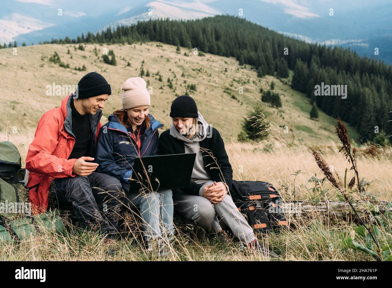 un gruppo di turisti tre persone un uomo e due donne mentre si rilassano in montagna si siedono su un albero caduto e guardano un computer portatile. Concetto di tecnologia Foto Stock