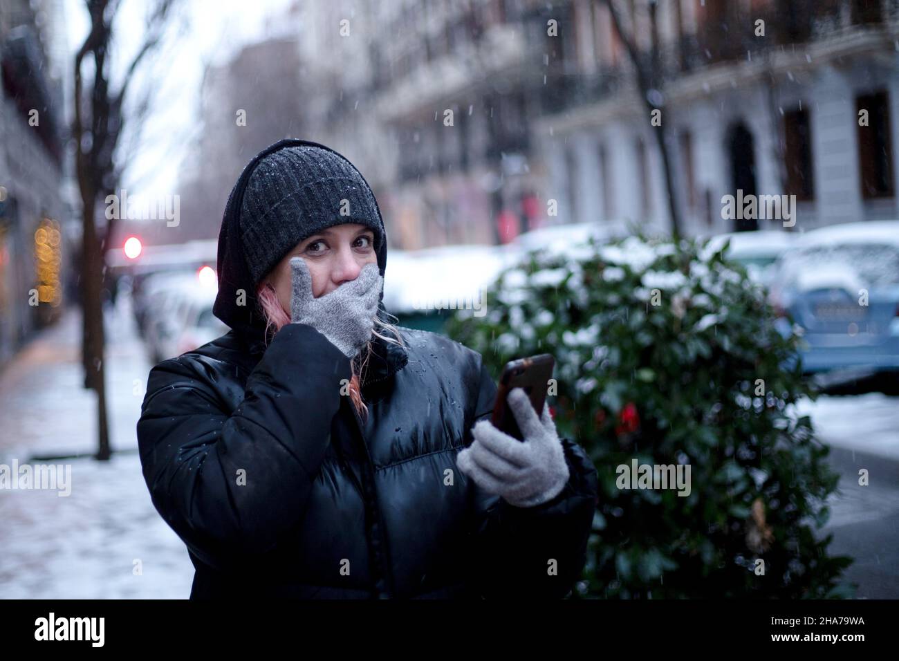 donna vestita come cappotto e cappello nella neve coprendo la bocca in sorpresa con cellulare in mano in città Foto Stock
