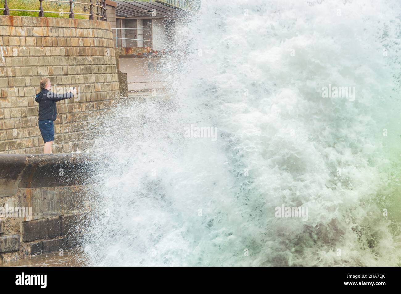 Uomo che fotografa mare tempestoso quasi inghiottito da onda gigante sul fronte mare di Scarborough Foto Stock