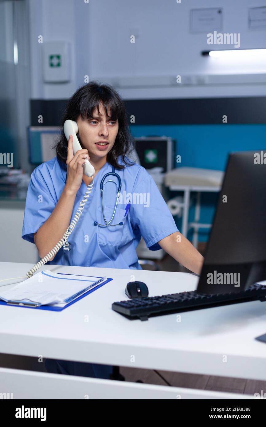 Assistente medico che parla al telefono per un appuntamento di check-up, lavorando tardi. Infermiere donna che utilizza il telefono fisso per la comunicazione remota online con il paziente presso l'armadietto sanitario. Foto Stock