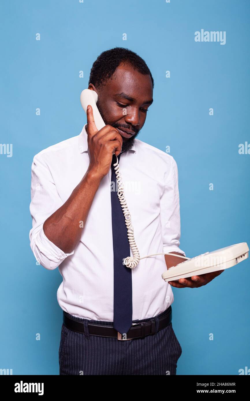 Tranquillo uomo d'affari che tiene recidiva per l'orecchio in piedi in su parlando sul telefono del cavo della linea fissa che ascolta la persona che parla all'altra estremità della linea. Dipendente in camicia bianca con telefono oldschool. Foto Stock