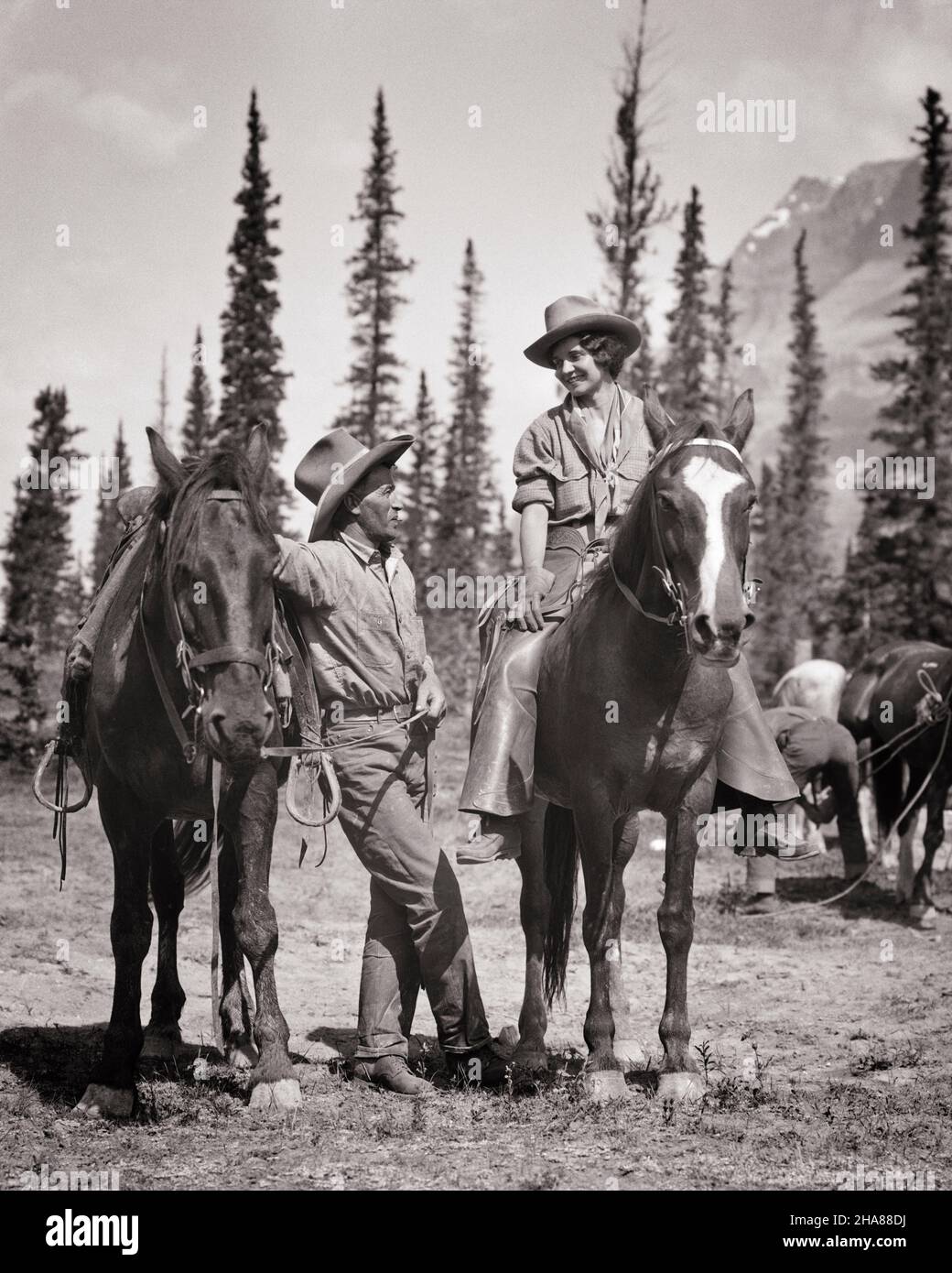 19S 1930S UOMO PIÙ ANZIANO RANCH MANO COWBOY IN PIEDI A CAVALLO PARLANDO CON LA GIOVANE DONNA MONTATA COWGIRL A CAVALLO AL BEST WESTERN DUDE RANCH - H4008 HAR001 HARS BELLEZZA VECCHIO
