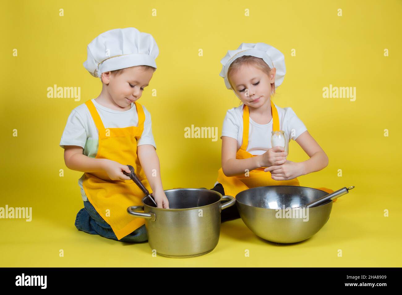 Bambini caucasici che giocano chef, bambini in grembiule e cappello dello chef seduti sul pavimento con una ciotola e sussurrare su sfondo giallo Foto Stock