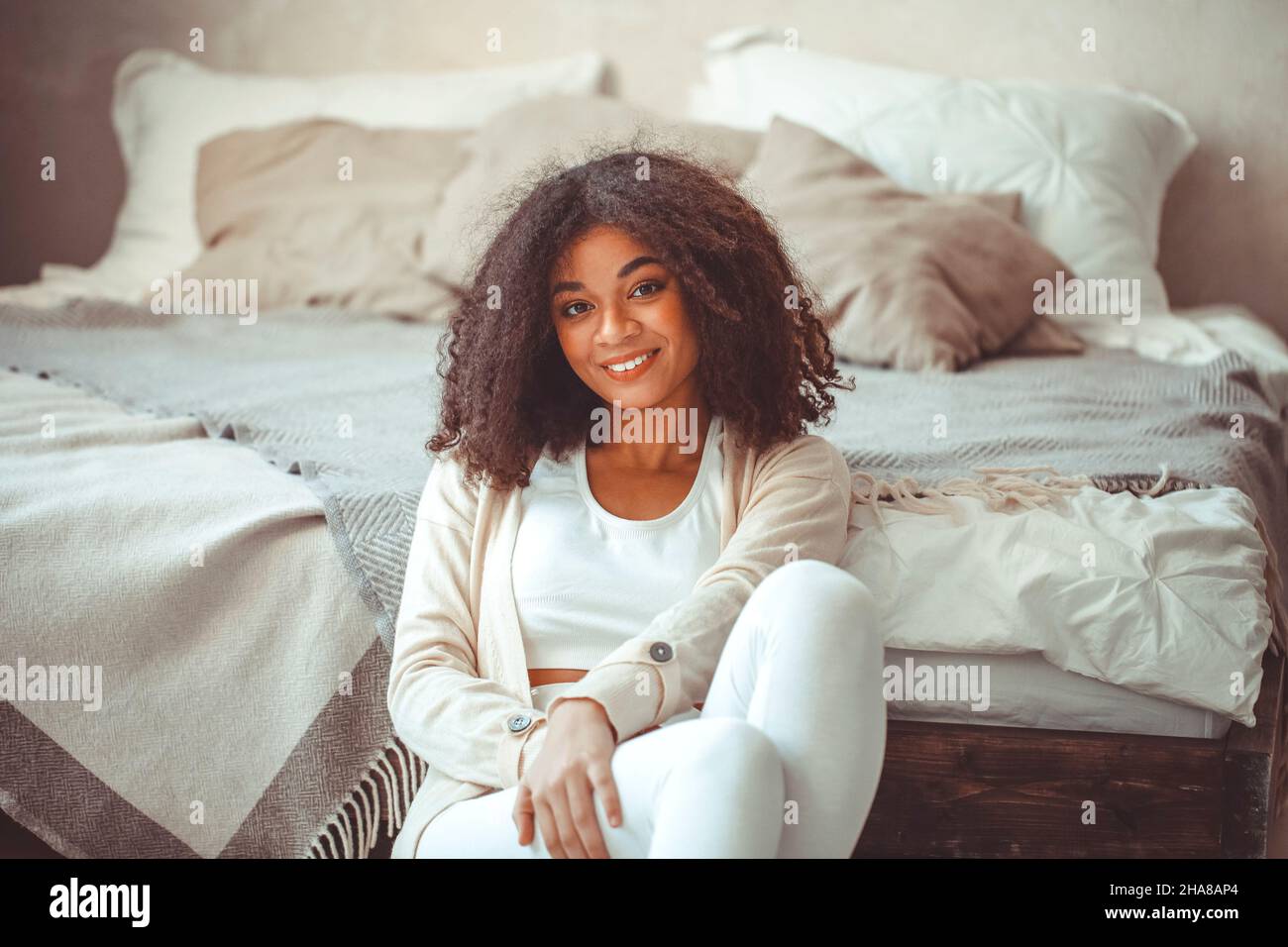 Allegra ragazza afroamericana affascinante con capelli ricci in camera da letto, si siede sul pavimento a piedi di fatto letto matrimoniale, guardando la macchina fotografica con ampio smil Foto Stock