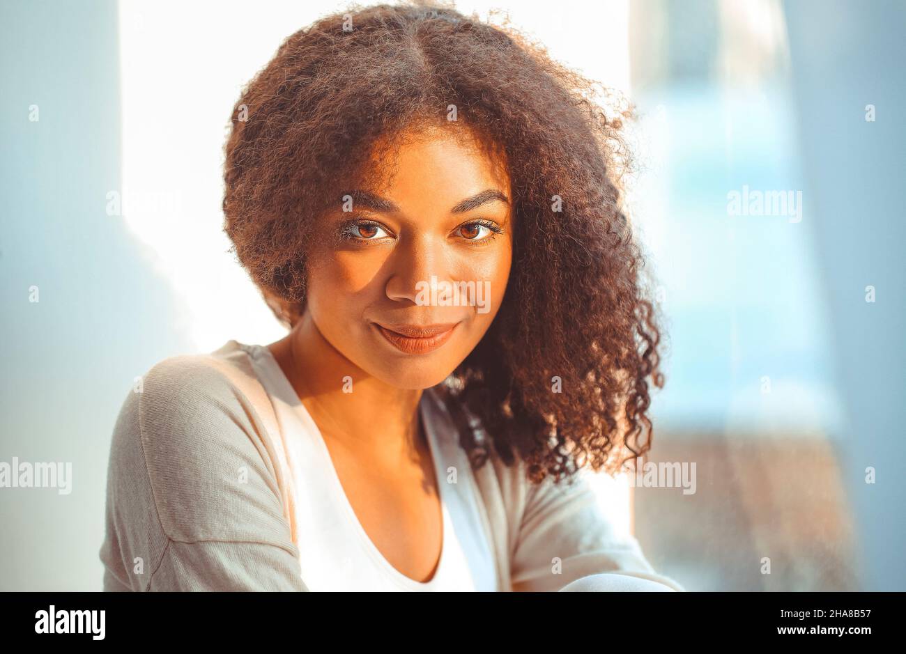 Felice African American ragazza etnia rilassato in abiti da casa seduti su davanzale sullo sfondo di tende che ondolano dal vento chiaro, felice afro Foto Stock