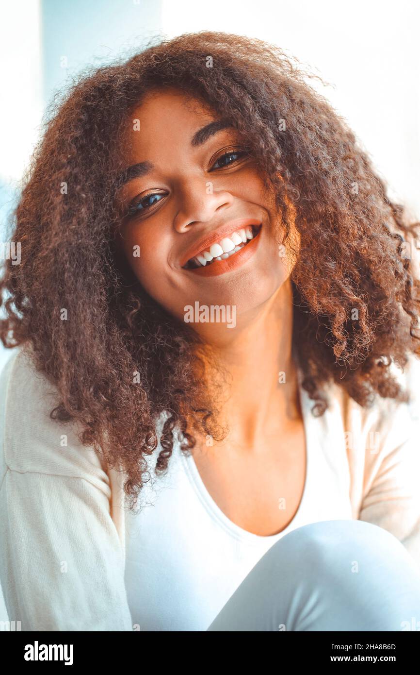 Felice African American ragazza etnia rilassato in abiti da casa seduti su davanzale sullo sfondo di tende che ondolano dal vento chiaro, felice afro Foto Stock