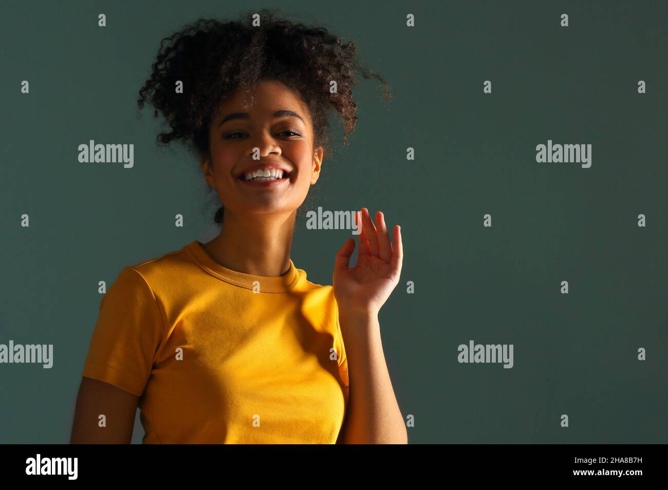 Bella ragazza di etnia afroamericana felice scuro con pelle in maglia gialla che alza la mano nel saluto mentre guarda in macchina fotografica con piacevole sorriso, p Foto Stock