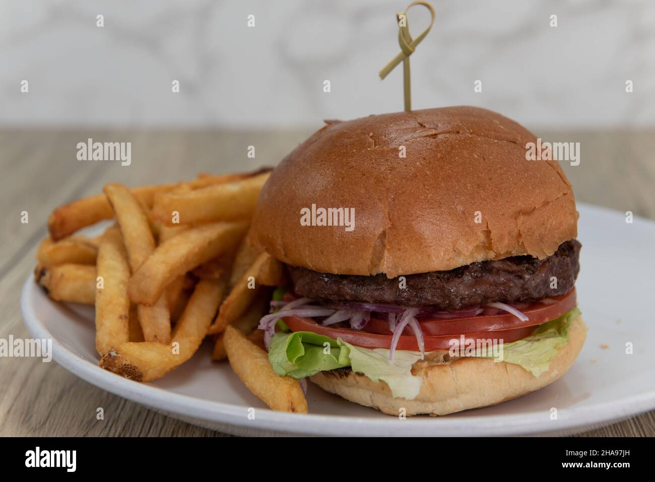 Presentazione artistica di un classico hamburger americano, con pomodoro a fette e cipolla su un panino di burro e servito con patatine fritte croccanti. Foto Stock