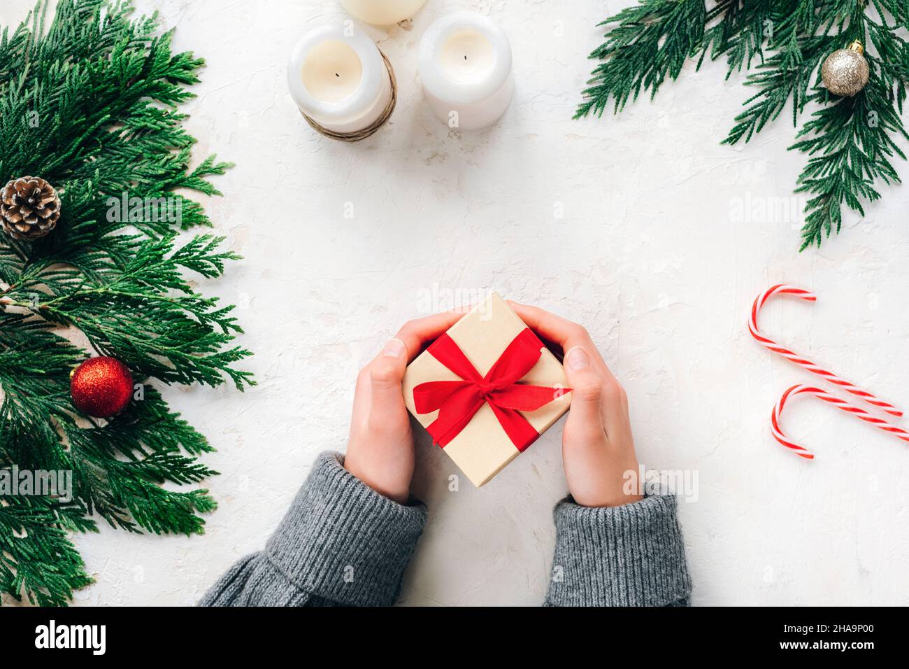 Le mani del bambino contengono la scatola minimalistica del regalo di Natale, decorata con il nastro rosso. Rami di abete, candele e caramelle su sfondo bianco testurizzato. Da a. Foto Stock