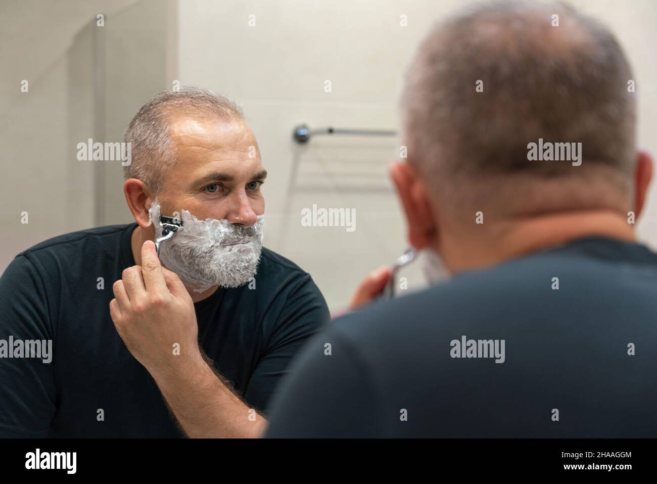 Uomo maturo dai capelli grigi che restyling la sua barba in casa utilizzando il rasoio Foto Stock