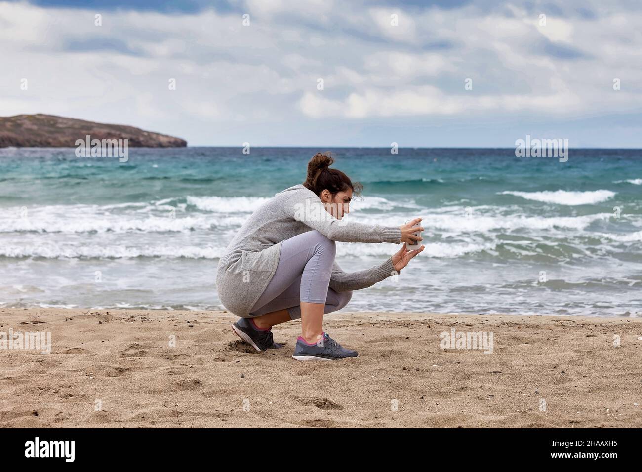 Donna che scatta una fotografia. Giovane fotografo che si piega sulla spiaggia sabbiosa scattando un colpo di mare con smartphone. Immagine stock, Foto Stock