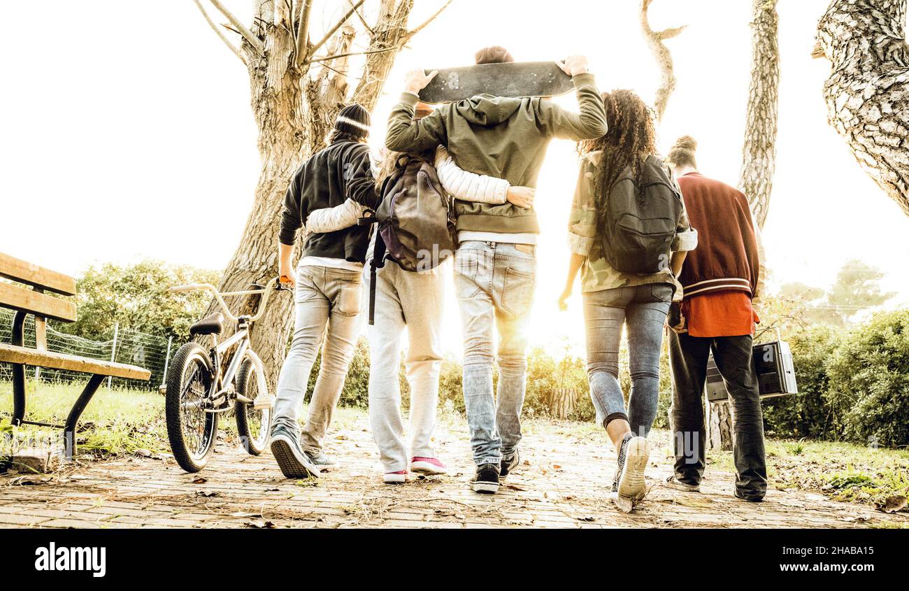 Gruppo di amici urbani che camminano nel parco di pattinare della città con retroilluminazione al tramonto - concetto di gioventù e amicizia con i giovani multirazziali che si divertono Foto Stock