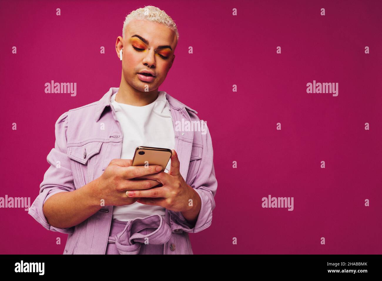 Sfogliare i podcast di tendenza. Giovane uomo elegante che utilizza uno smartphone indossando auricolari wireless in uno studio. Non conforme queer man in piedi da solo Foto Stock