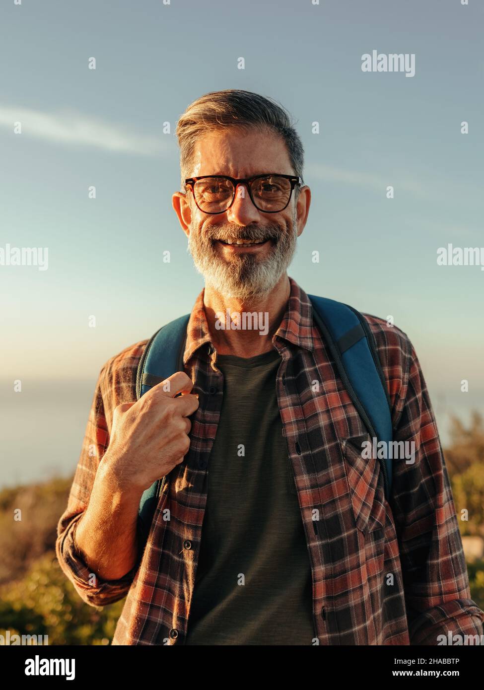 Hiker maturo e allegro sorridendo alla macchina fotografica mentre si alza da solo. Uomo maturo felice che porta uno zaino e si alza in cima a una collina. Zaino avventuroso Foto Stock