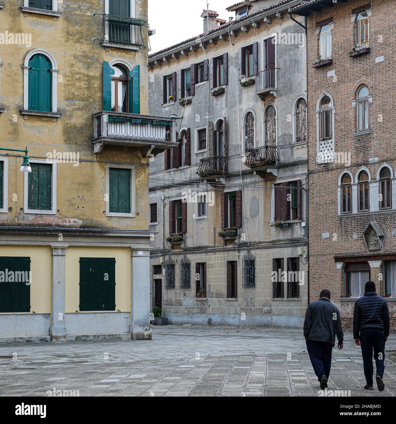 Due uomini neri stanno facendo una passeggiata in una piccola strada con case un po' buie a Venezia. È deserta a causa della pandemia di Corona. Foto Stock