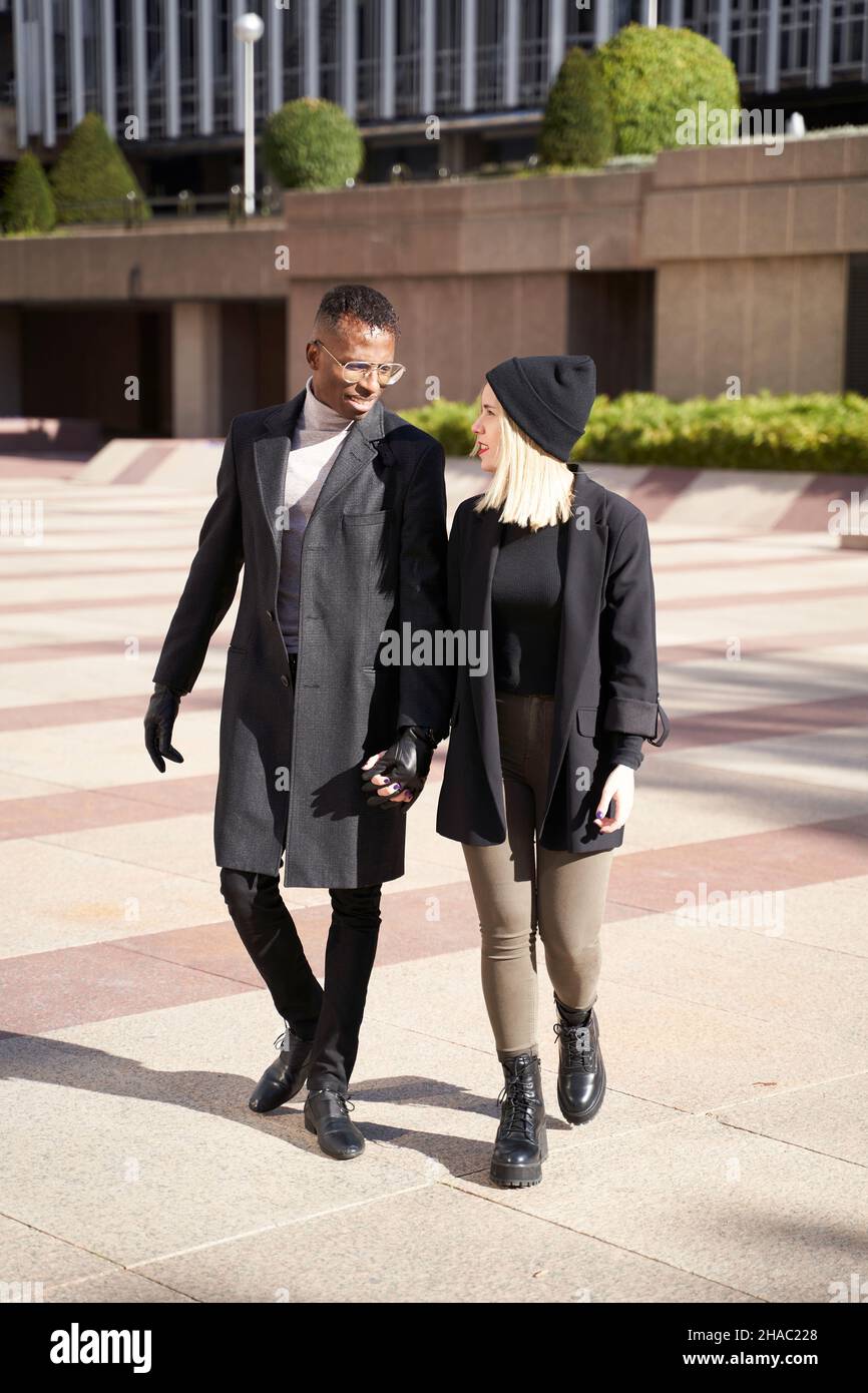 Uomo e donna multirazziale a corpo pieno in outerwear tenendo le mani e guardando l'un l'altro mentre passeggi sulla strada e parla Foto Stock