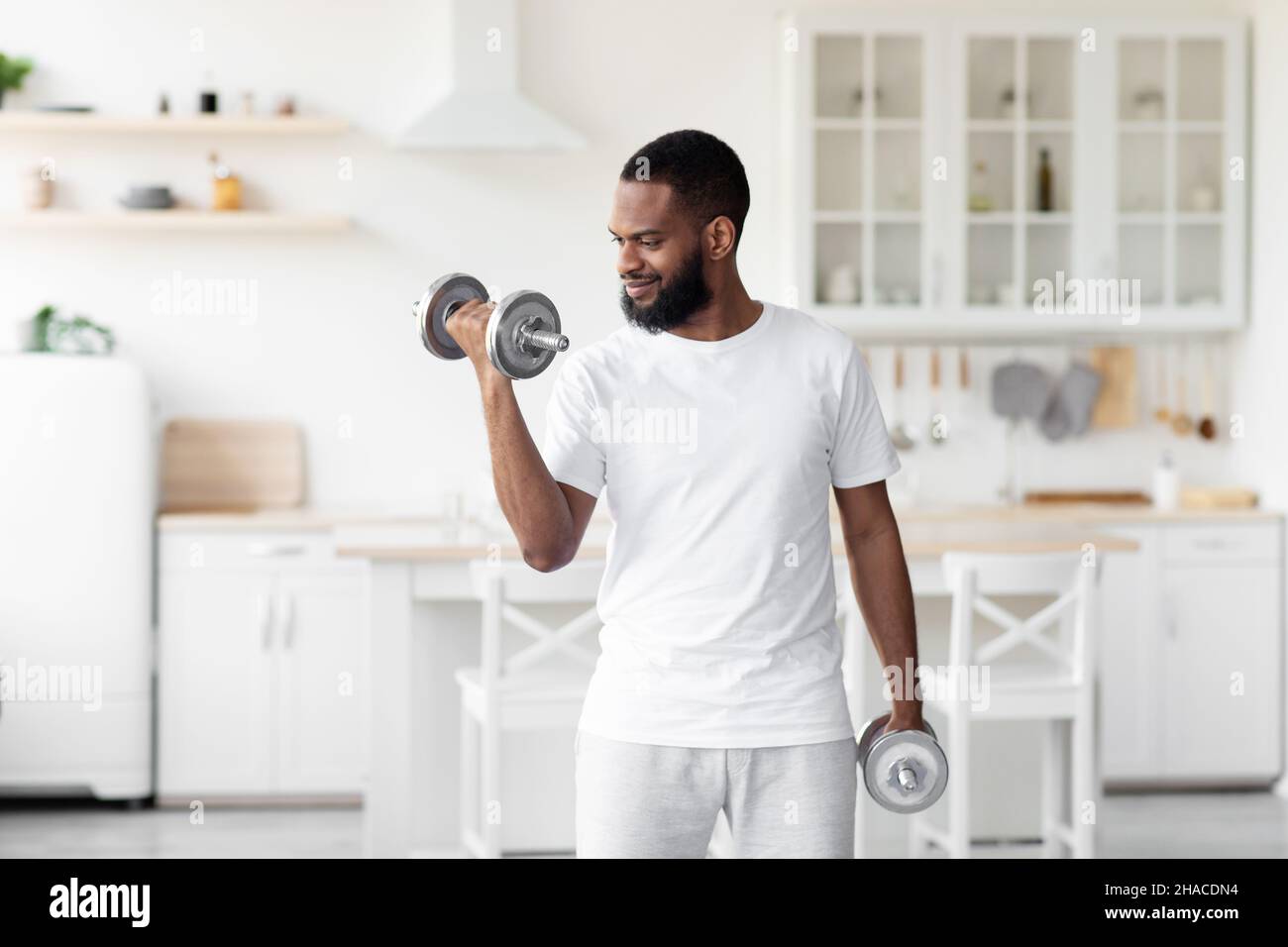 Sorridente millennial bel ported africano americano maschio atleta in abiti bianchi solleva manubelli in cucina minimalista Foto Stock