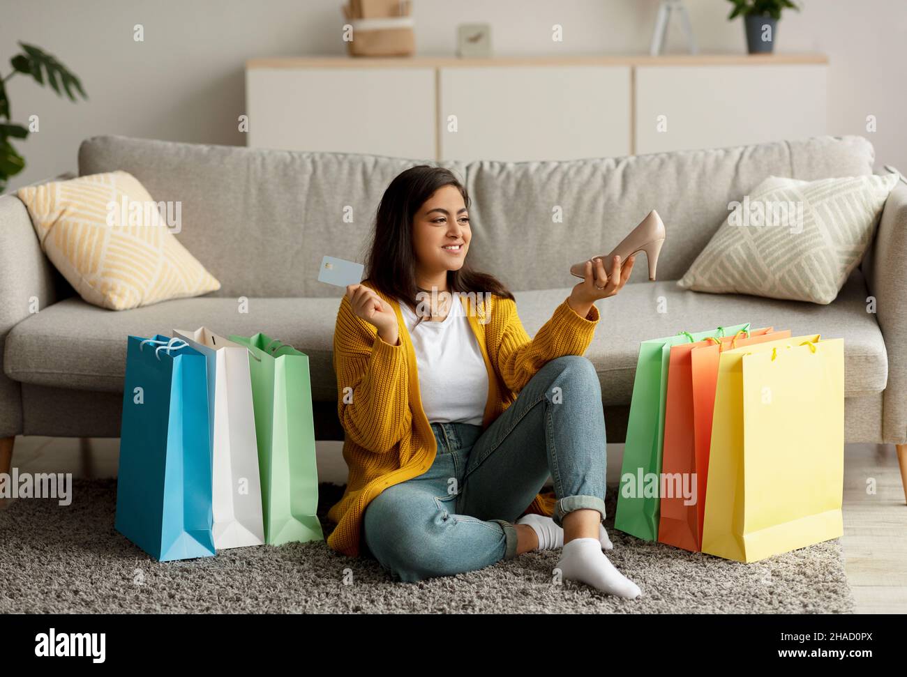 Pagamento facile, shopping remoto. Donna araba felice che tiene carta di credito e nuova scarpa, seduta sul pavimento tra borse shopper Foto Stock